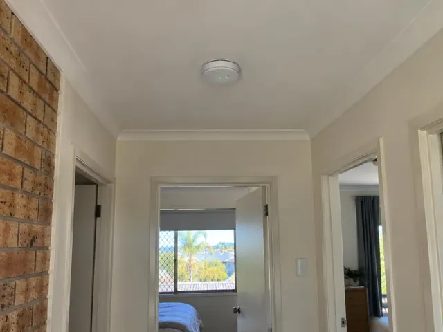 Ceiling-mounted smoke alarm in a hallway with doorways and brick wall in Bundall